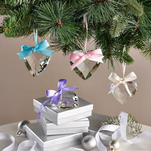 Four glass heart-shaped ornaments with delicate ribbons are suspended from a Christmas tree branch, accompanied by gift boxes tied with complementary satin ribbons below.