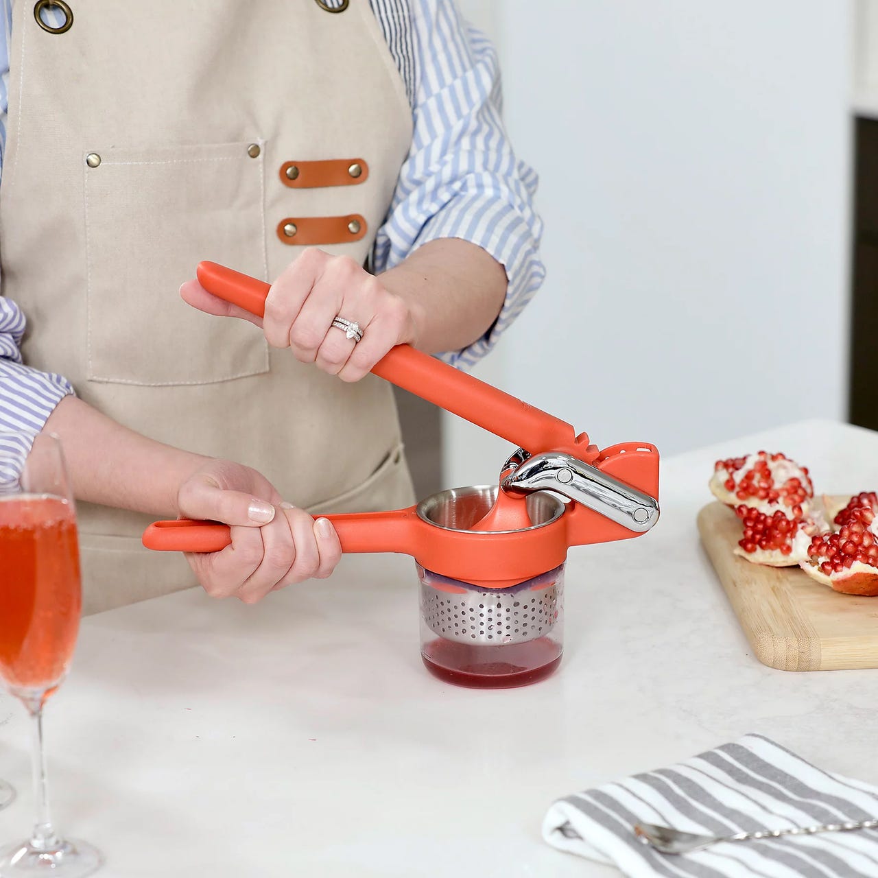 A person uses a red handheld citrus press juicer to extract juice into a container. A pomegranate is partially prepared on a cutting board in the background.
