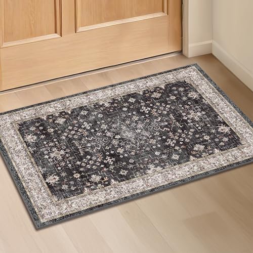 A rectangular patterned rug with a dark central design and a contrasting light border is placed in front of a wooden door on a light-colored floor.