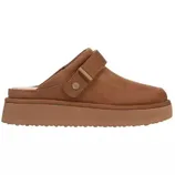 A tan slip-on mule with a thick sole and adjustable strap, featuring a subtle stitched design and a small metal accent.