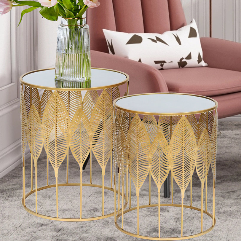 Set of 2 gold-toned nesting end tables with a decorative leaf pattern and mirrored tops, shown next to a pink chair and pillow.