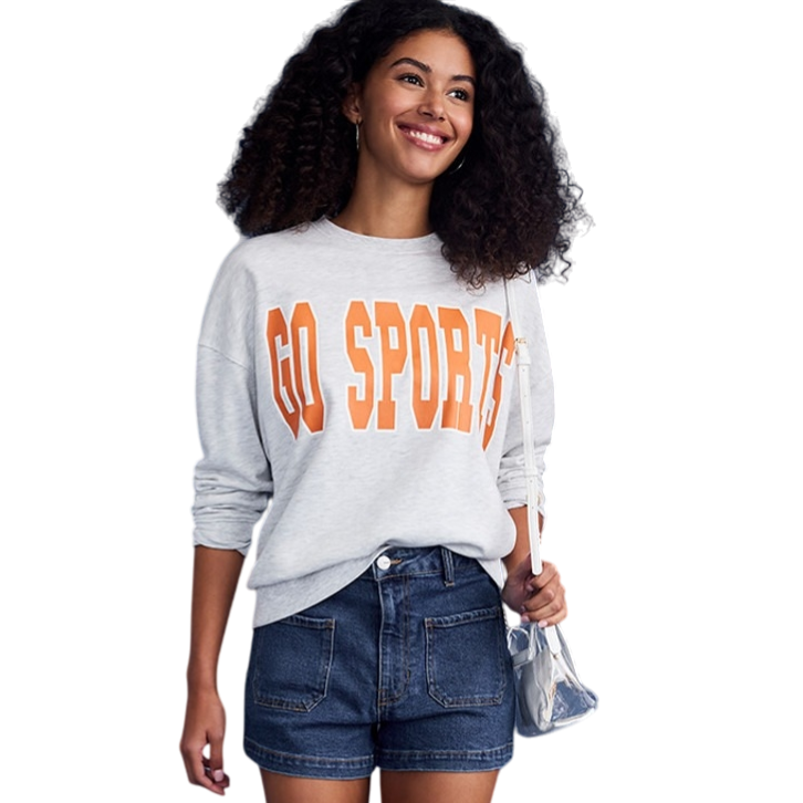 A person is wearing a gray sweatshirt with \“GO SPORTS\“ in orange lettering, denim shorts, and beige sneakers. They are holding a clear crossbody bag.