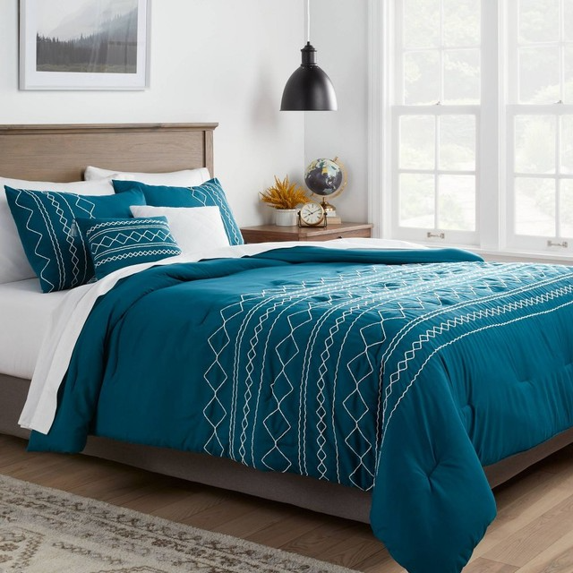 A teal 5-piece queen comforter set featuring white geometric embroidery, including a comforter, two pillow shams, and two decorative pillows. The set is displayed on a bed with a modern wooden headboard in a bright, well-lit room.