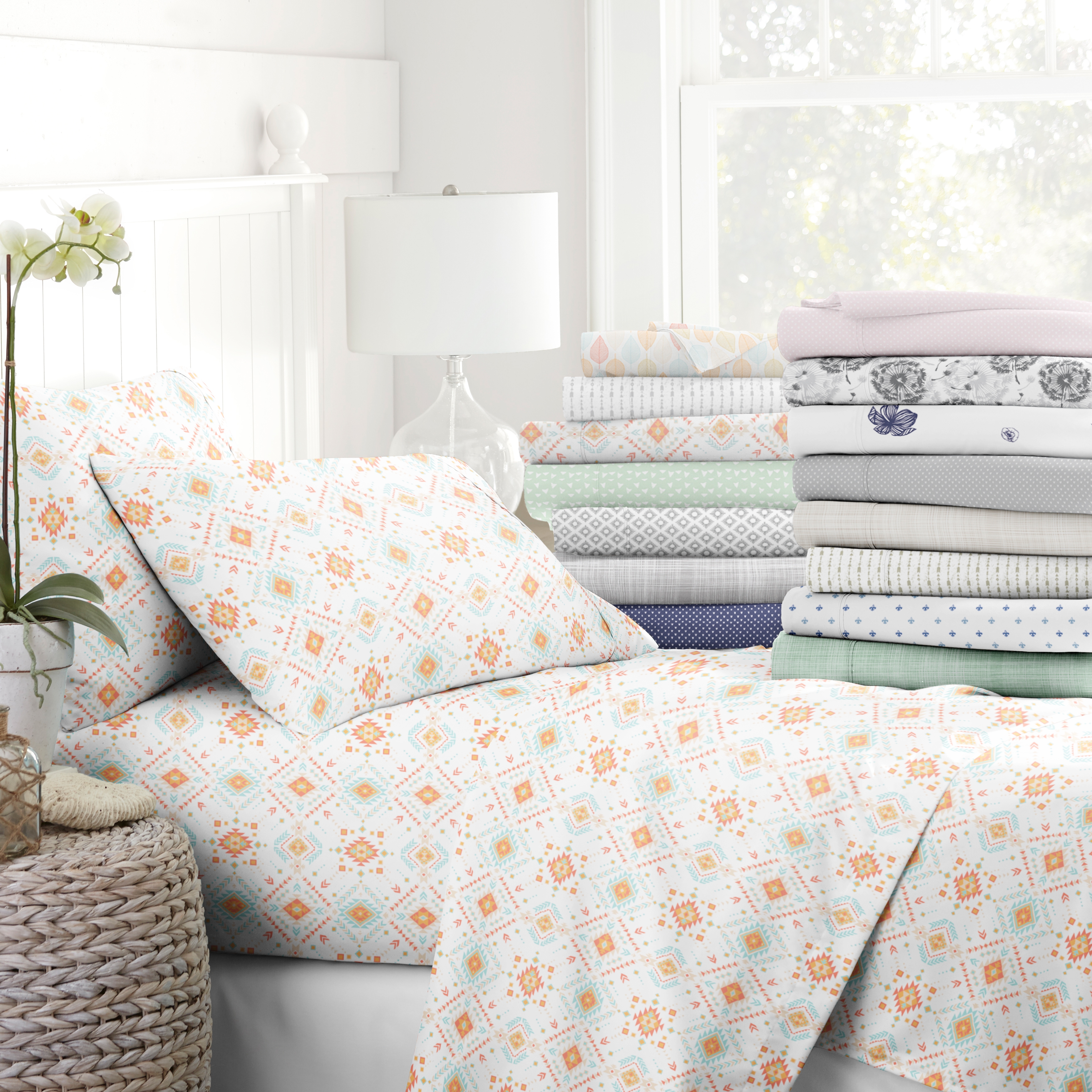 A variety of patterned bed sheets and pillowcases displayed on a bed and stacked nearby, with a lamp and plant accenting the scene.