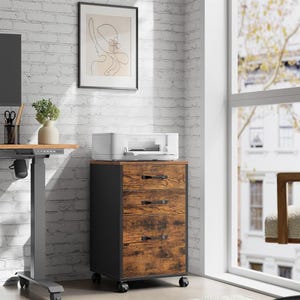 A modern home office setup with a white brick wall, featuring a standing desk, a printer on a wooden filing cabinet with wheels, and a framed abstract artwork.