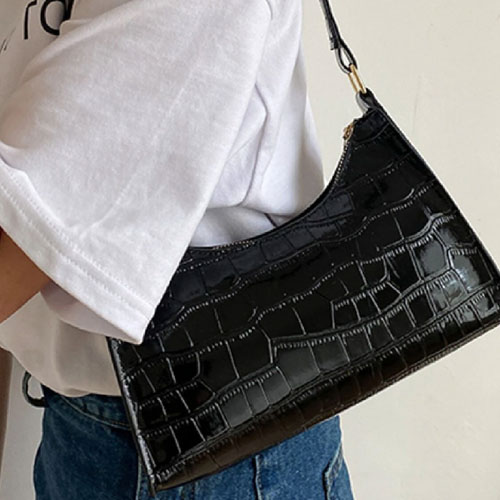 A person is wearing a white t-shirt and blue jeans, holding a black, crocodile pattern shoulder bag.