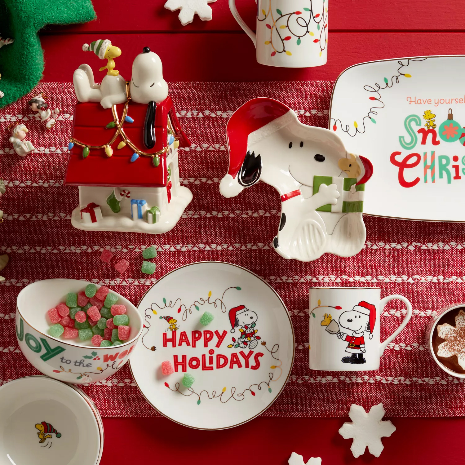 Festive tableware featuring a Christmas theme with Snoopy and Woodstock, including plates, a mug, a cookie jar shaped like Snoopy's doghouse, and holiday treats on a red cloth background.