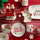 Festive tableware featuring a Christmas theme with Snoopy and Woodstock, including plates, a mug, a cookie jar shaped like Snoopy's doghouse, and holiday treats on a red cloth background.
