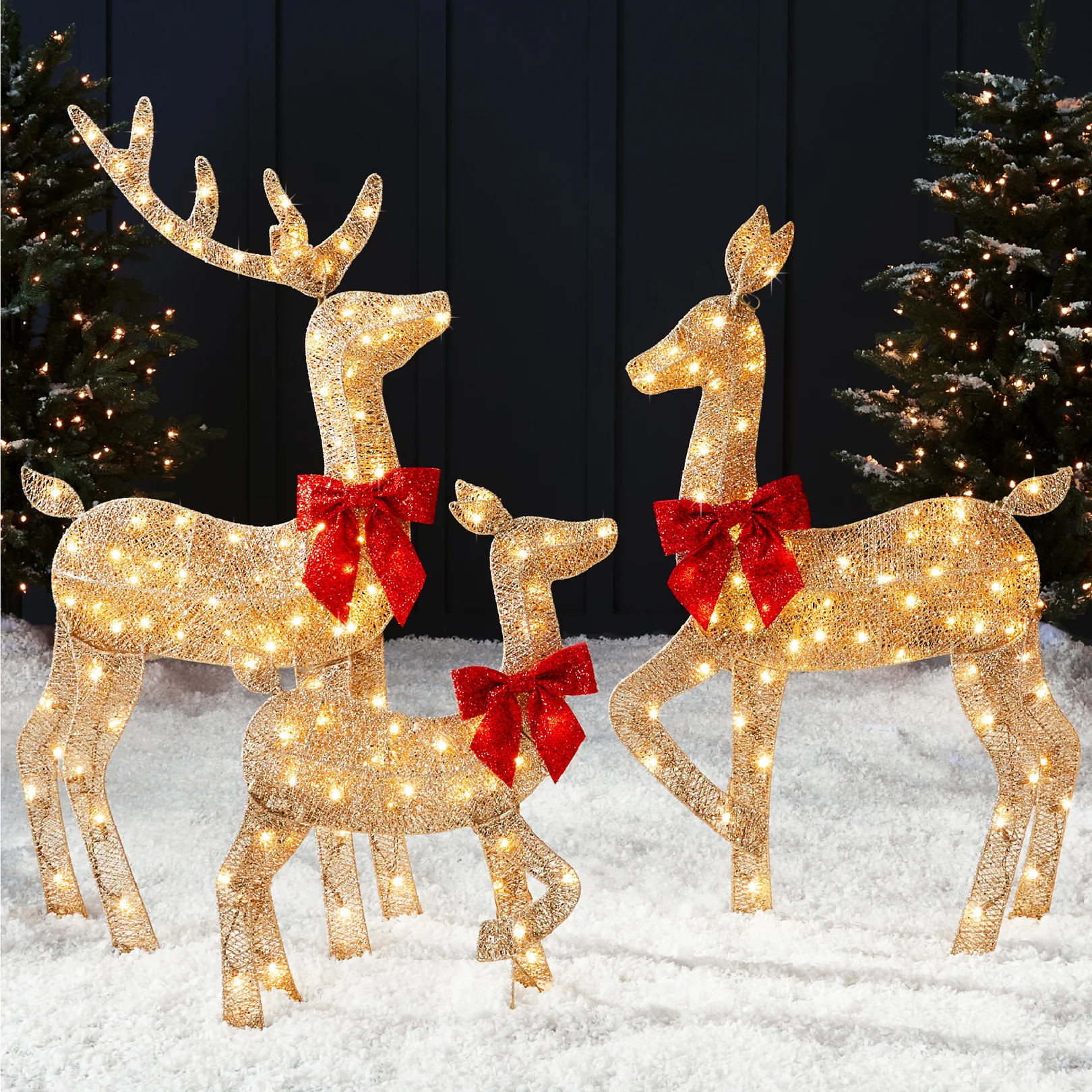 A set of three illuminated reindeer sculptures is displayed, adorned with red bows and lights, standing on artificial snow with decorated trees in the background.