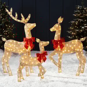 A set of three illuminated reindeer sculptures is displayed, adorned with red bows and lights, standing on artificial snow with decorated trees in the background.