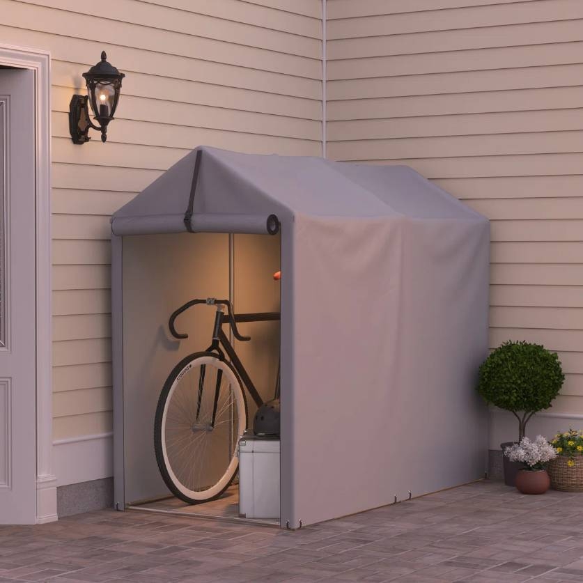 A compact, gray portable storage shed is attached to a house wall, housing a bicycle, helmet, and a white box. It's constructed with a fabric covering and has a peaked roof design.