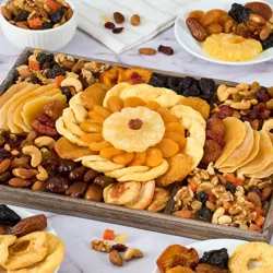 A wooden tray displays an assortment of dried fruits and nuts, including apricots, pineapples, apples, dates, walnuts, almonds, cashews, and mixed berries. Nearby, small plates hold similar assortments.
