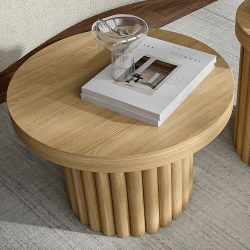 A round wooden coffee table with a ribbed cylindrical base is topped with a stack of magazines and a transparent glass mug.