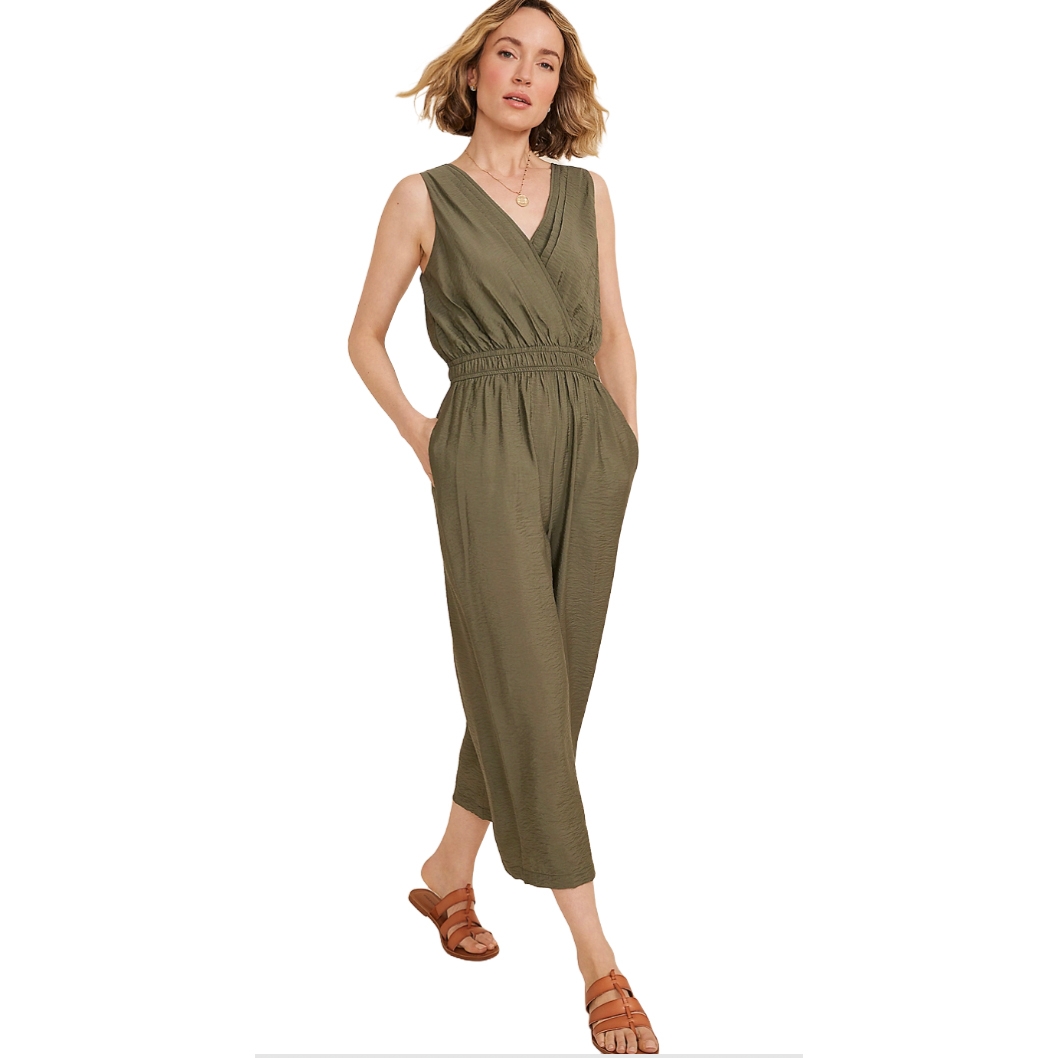Olive green sleeveless jumpsuit with a V-neck wrap front, elastic waistband, and cropped wide legs. Paired with brown flat sandals.