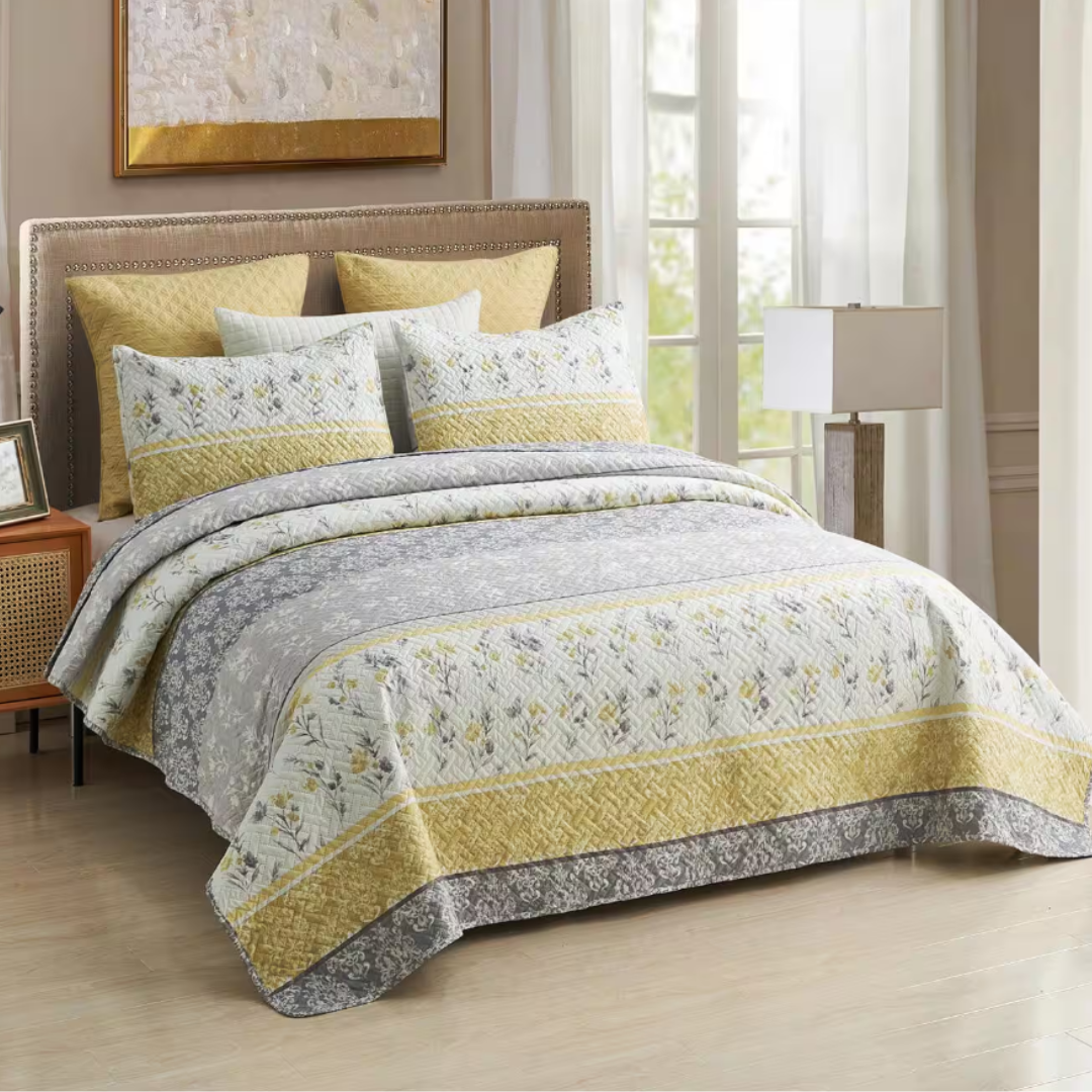 A bed features a quilt set with a floral and geometric pattern in gray and yellow tones, complemented by matching pillows. The room includes a lamp, a wooden nightstand, and a large window with sheer curtains.