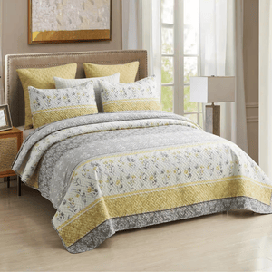 A bed features a quilt set with a floral and geometric pattern in gray and yellow tones, complemented by matching pillows. The room includes a lamp, a wooden nightstand, and a large window with sheer curtains.