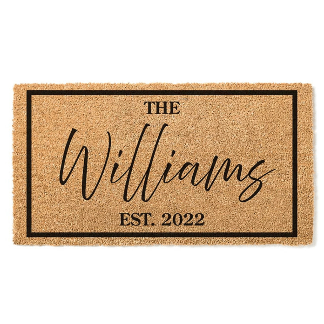 A rectangular doormat features \“The Williams\“ in large cursive with \“EST. 2022\“ below, all set within a simple black border on a natural coir background.