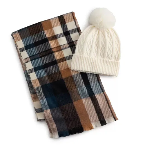 A cream cable-knit beanie with a fluffy pom-pom is paired with a plaid scarf in brown, black, and blue tones.