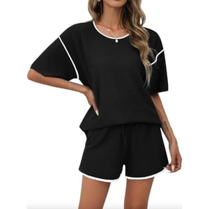A woman is wearing a black pajama set with short sleeves and shorts, featuring white trim details around the sleeves, neckline, and hem. She has long, wavy hair and a necklace.