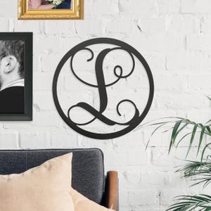 A black metal wall art piece with a decorative letter \“L\“ in a circular design hangs on a white brick wall, alongside framed photos, above a gray couch with beige cushions and next to a plant.