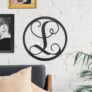 A black metal wall art piece with a decorative letter \“L\“ in a circular design hangs on a white brick wall, alongside framed photos, above a gray couch with beige cushions and next to a plant.