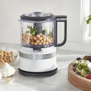 KitchenAid food processor with chickpeas and herbs inside, placed on a kitchen counter next to a salad.