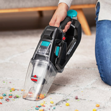 A handheld vacuum cleaner is being used to pick up colorful cereal pieces from a carpet.