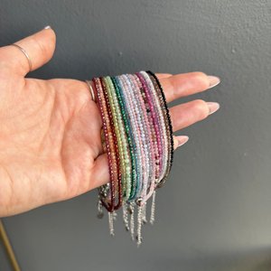 A collection of multicolored beaded anklets made from various crystal gemstones, displayed in a hand. Each anklet features small, round beads and some have dangling silver chain accents.