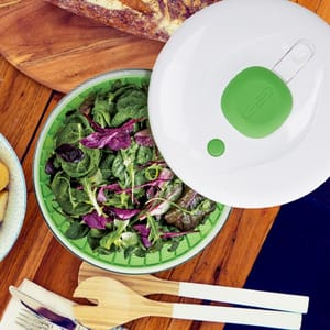 A green and white salad spinner next to a bowl of mixed leafy greens and kitchen utensils on a wooden surface.