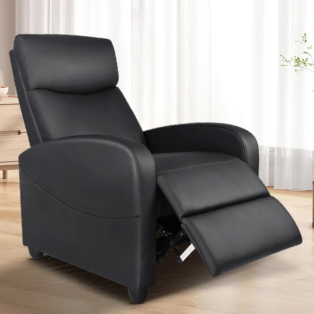 A black recliner chair with a cushioned back, armrests, and an extended footrest positioned in a bright room with wooden flooring.