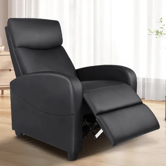 A black recliner chair with a cushioned back, armrests, and an extended footrest positioned in a bright room with wooden flooring.