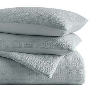 A stack of textured light blue bedding, including pillows and a quilt or blanket with a woven, dotted pattern.