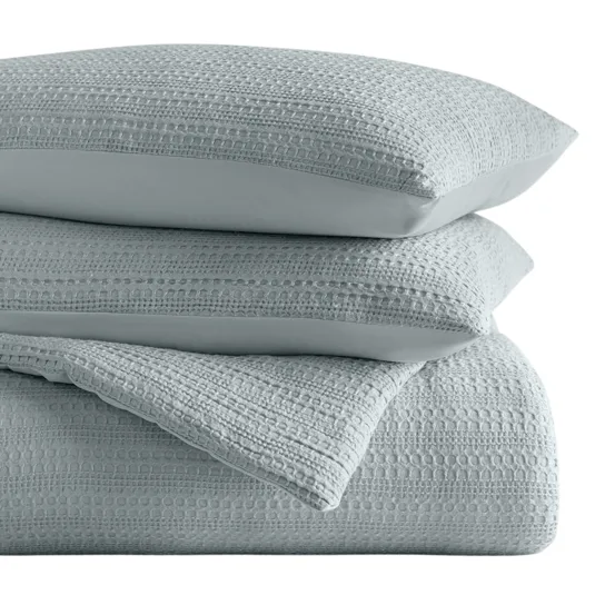 A stack of textured light blue bedding, including pillows and a quilt or blanket with a woven, dotted pattern.