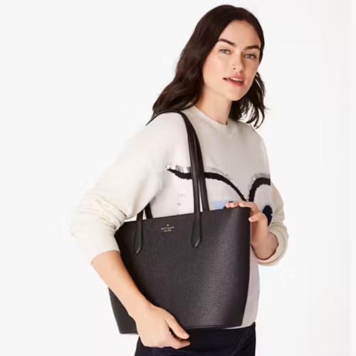 A woman holding a black tote bag and wearing a white sweatshirt with black abstract design.