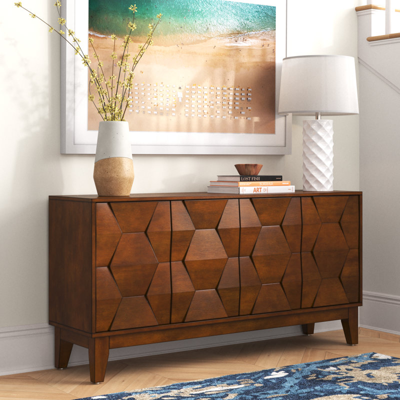 A wooden sideboard with geometric patterns stands beneath a framed beach picture, alongside a table lamp, vase with twigs, and books.