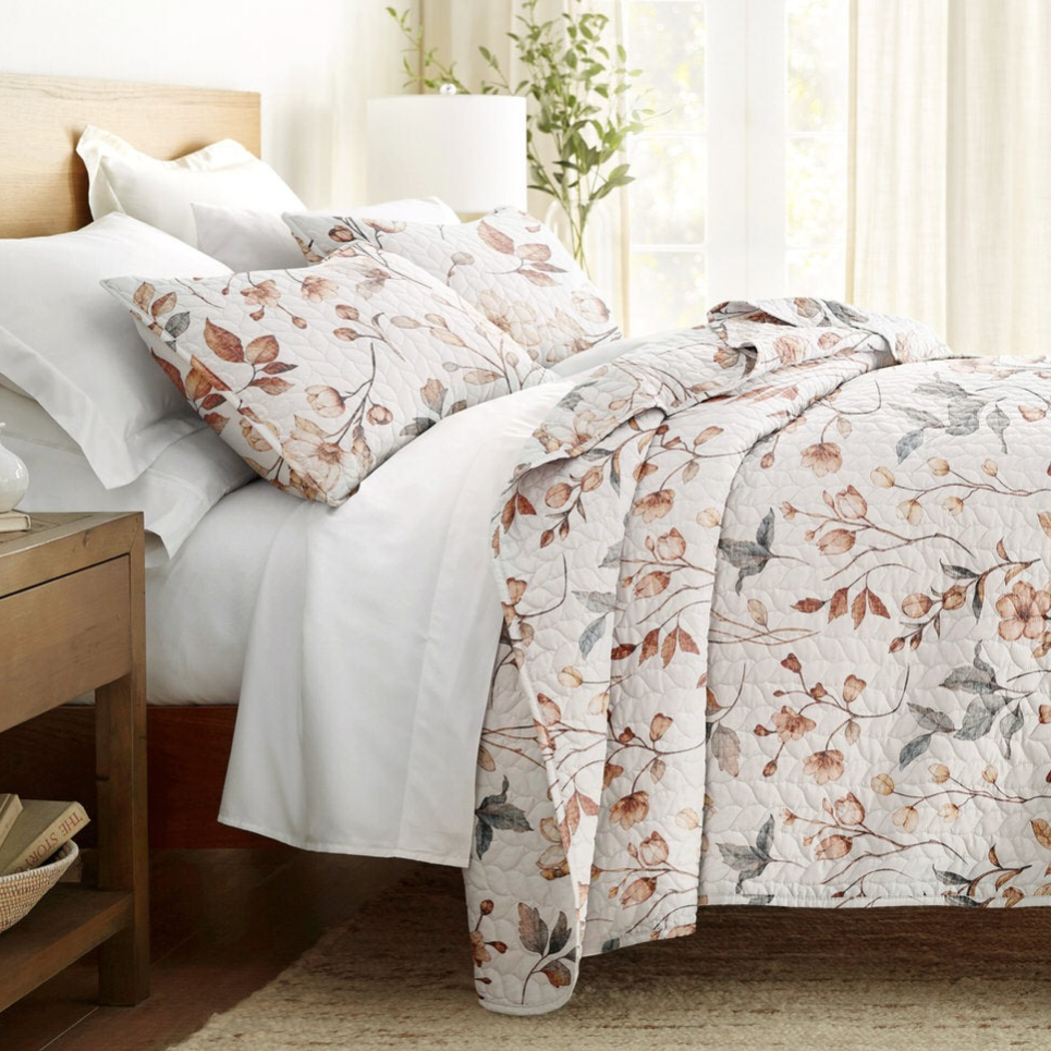 A bed is adorned with a quilt and pillowcases featuring a floral pattern with earthy tones, accompanied by white sheets. A wooden bedside table is nearby, holding books and a lamp.