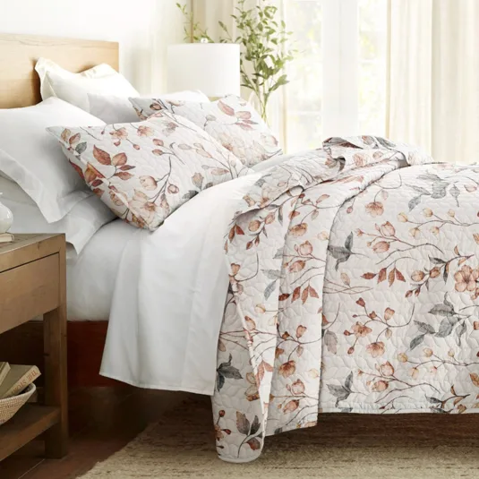 A bed is adorned with a quilt and pillowcases featuring a floral pattern with earthy tones, accompanied by white sheets. A wooden bedside table is nearby, holding books and a lamp.