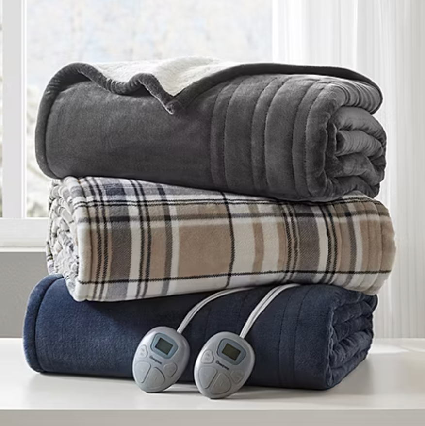 Three folded electric blankets, one gray, one plaid, and one navy, with two digital controllers, are stacked on a white surface in front of a window.