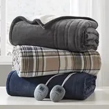 Three folded electric blankets, one gray, one plaid, and one navy, with two digital controllers, are stacked on a white surface in front of a window.