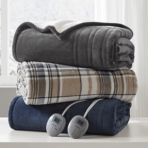 Three folded electric blankets, one gray, one plaid, and one navy, with two digital controllers, are stacked on a white surface in front of a window.