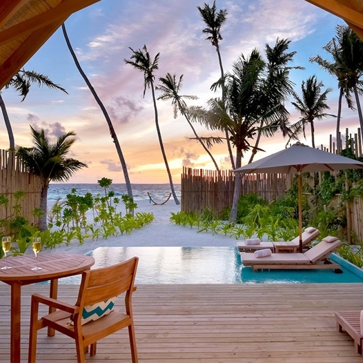 A beachfront deck features a small pool, two sun loungers with an umbrella, and a table with two chairs and wine glasses, overlooking the ocean and palm trees at sunset.