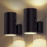 Two black wall-mounted cylindrical LED wall lights illuminate both upwards and downwards, installed above electrical outlets.