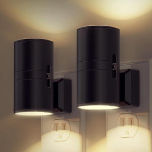 Two black wall-mounted cylindrical LED wall lights illuminate both upwards and downwards, installed above electrical outlets.