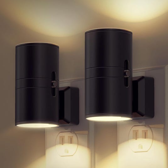 Two black wall-mounted cylindrical LED wall lights illuminate both upwards and downwards, installed above electrical outlets.