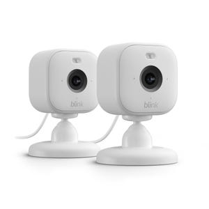 The 2pk Blink Mini Cameras are compact, white indoor security cameras designed for easy installation, featuring HD video, motion detection, and two-way audio. They rest on adjustable stands for flexible positioning and require a power source via cable.