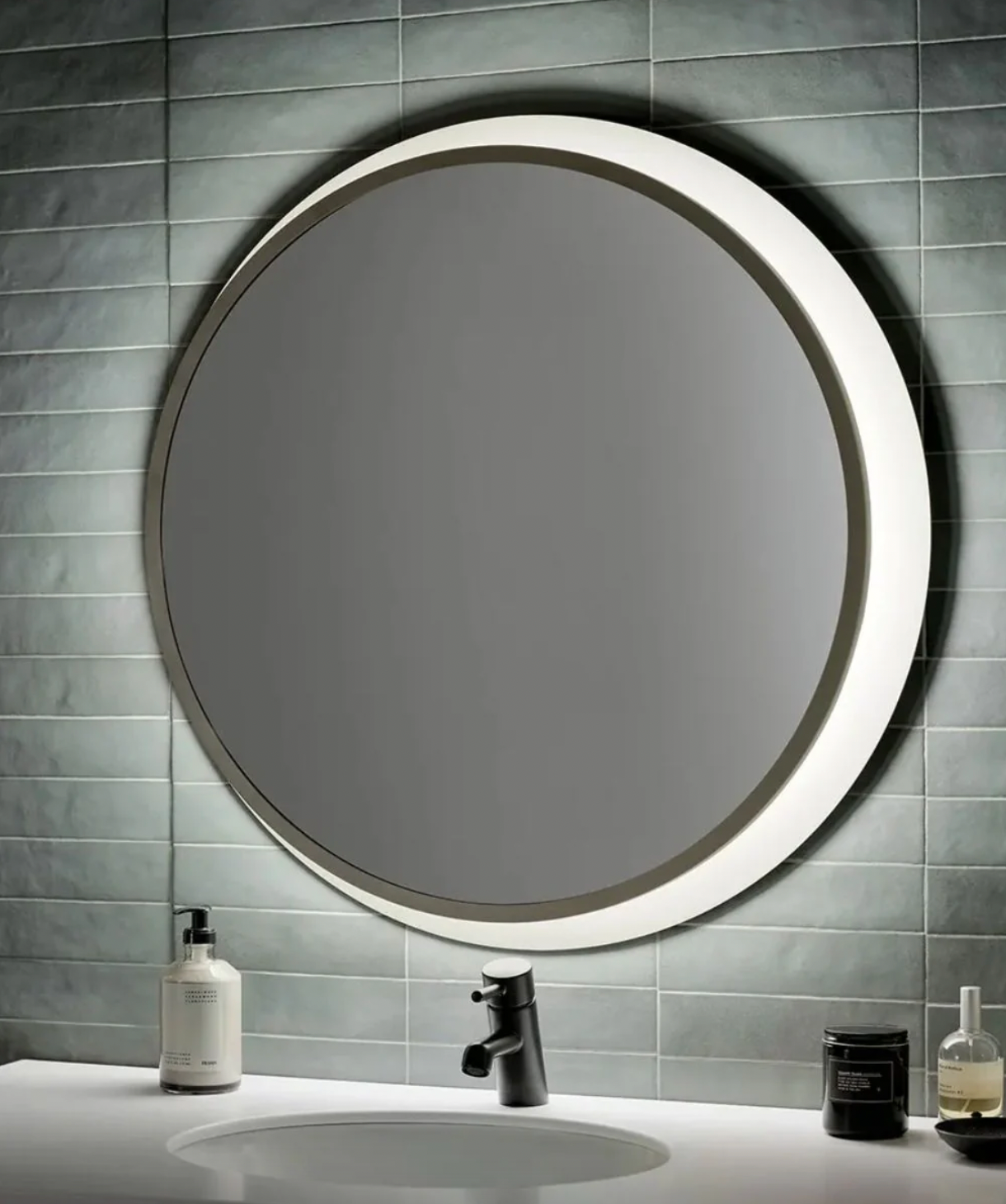 A round, 30-inch backlit LED wall mirror is mounted on a tiled wall above a white sink, featuring modern black faucet and adjacent with lotion and candles.