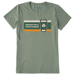 Green T-shirt featuring the text \“Drinks Well with Others\“ alongside a stylized beer glass and shamrock design, with orange and white accents. Brand: Life is Good.
