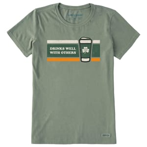 Green T-shirt featuring the text \“Drinks Well with Others\“ alongside a stylized beer glass and shamrock design, with orange and white accents. Brand: Life is Good.