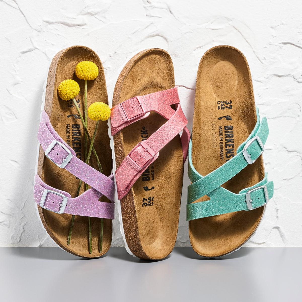 Two pairs of sparkly sandals in pink and mint green with adjustable straps, displayed with yellow pompom flowers between them.
