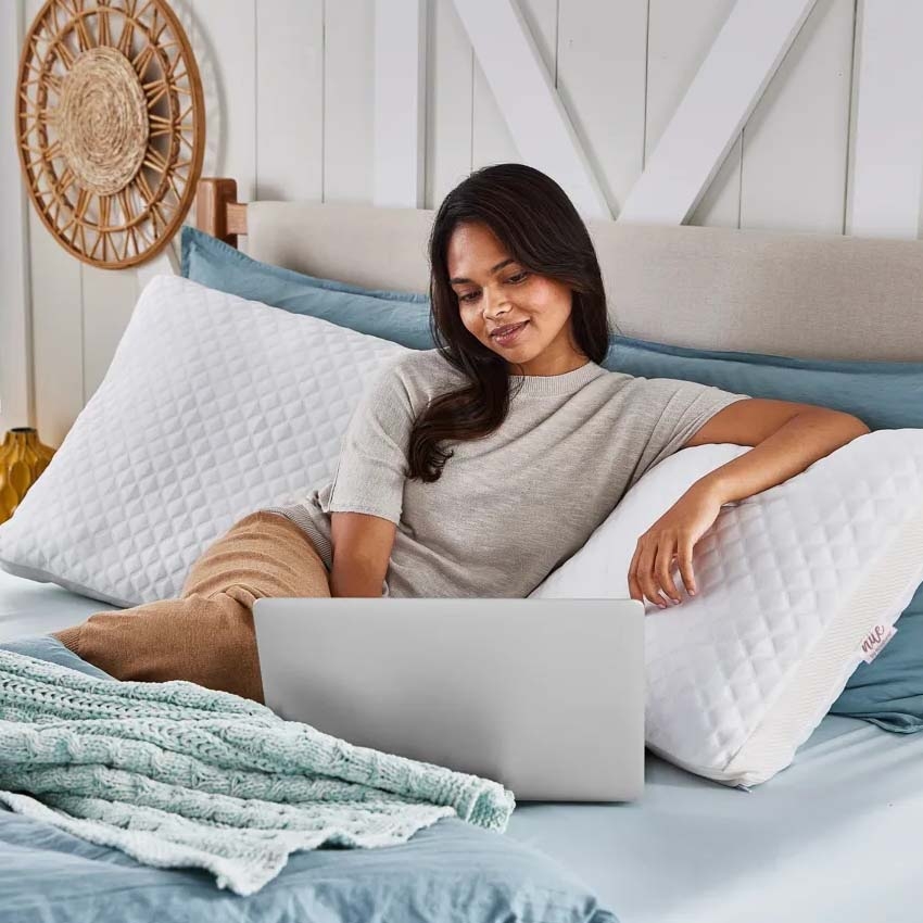 White quilted body pillows with a textured cover are placed horizontally behind a person sitting on a bed. They are next to light blue pillows and a knitted teal throw blanket.